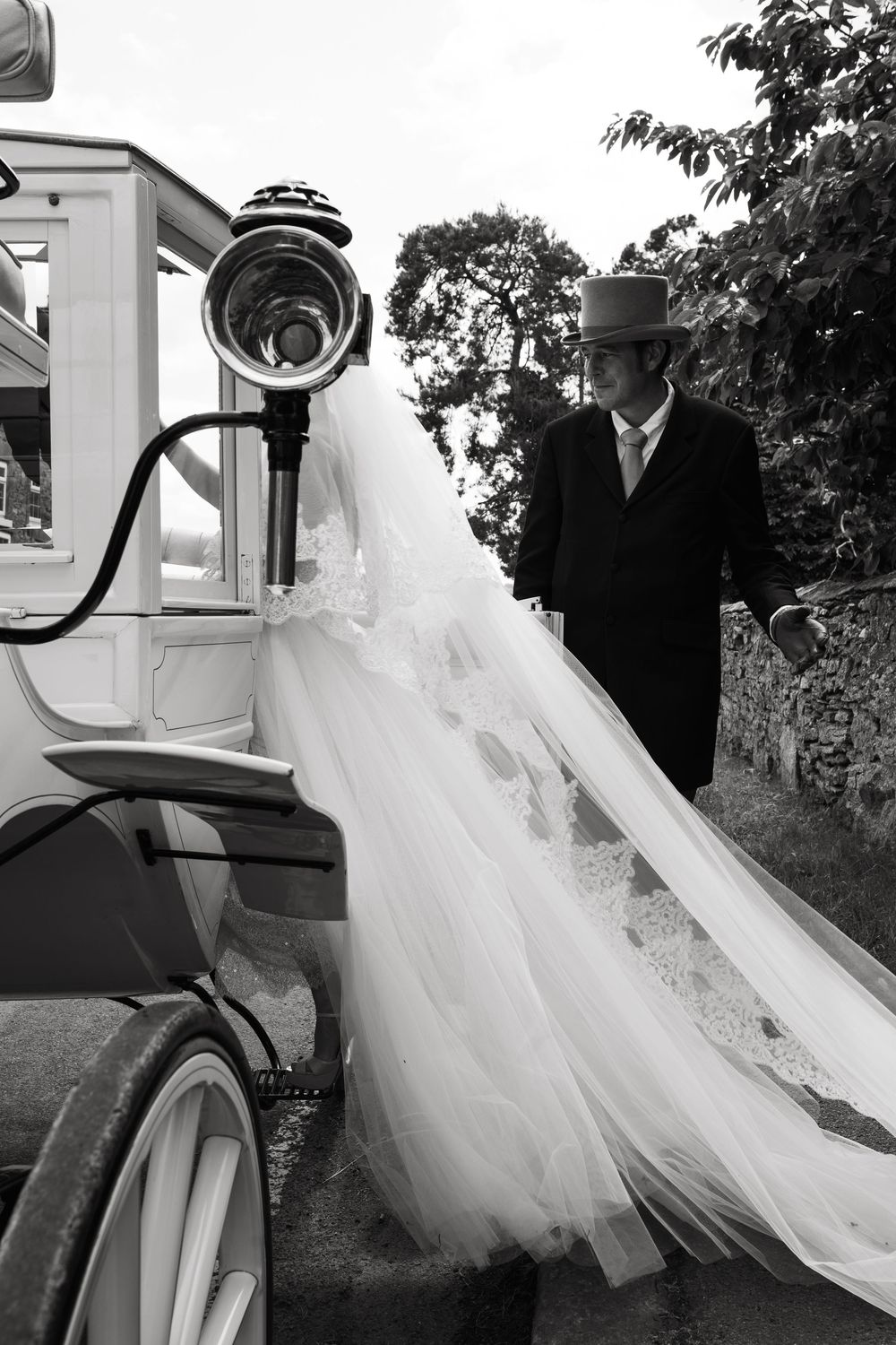 Vintage-style black and white photo of wedding dress flowing from classic car.