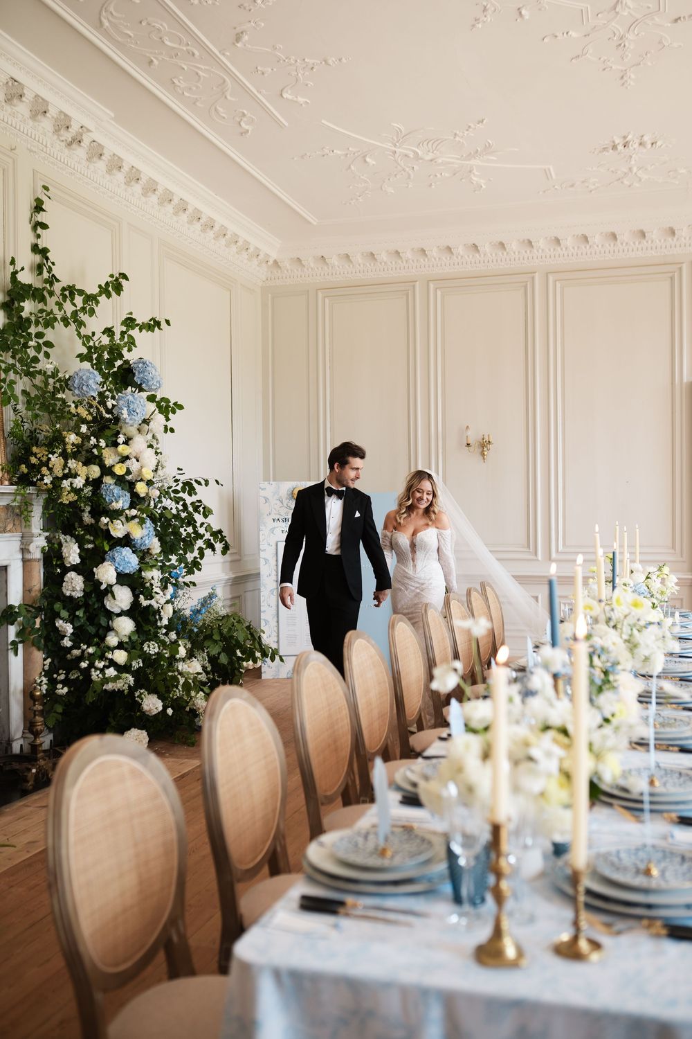 Newlyweds walk along an elegantly set dining table in a luxurious white room.