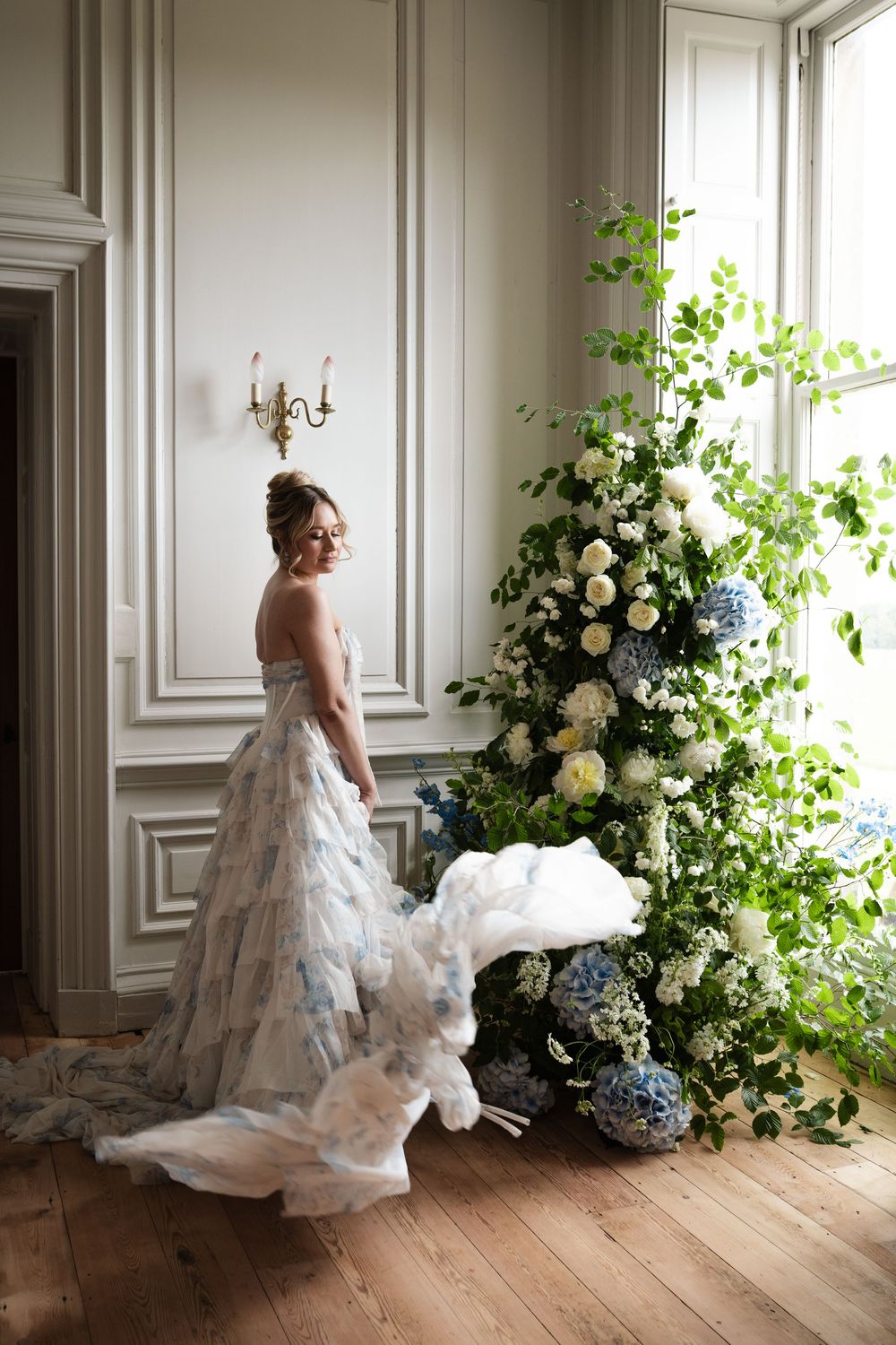 A bride in a layered white gown poses beside luxurious floral arrangements.