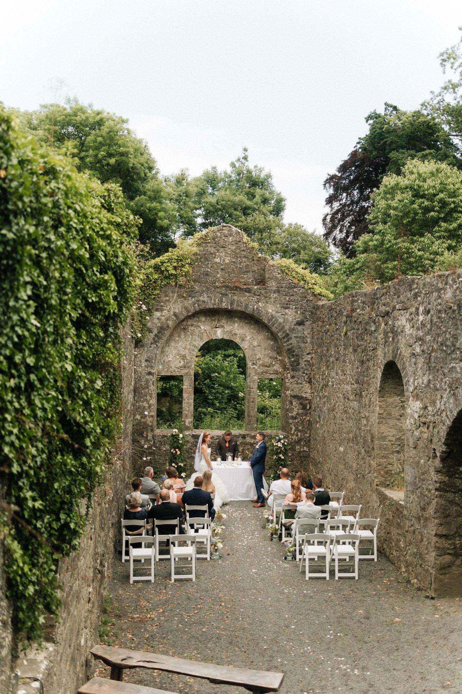 An intimate wedding ceremony takes place in the romantic ruins of an old stone castle with ivy-covered walls and white chairs.