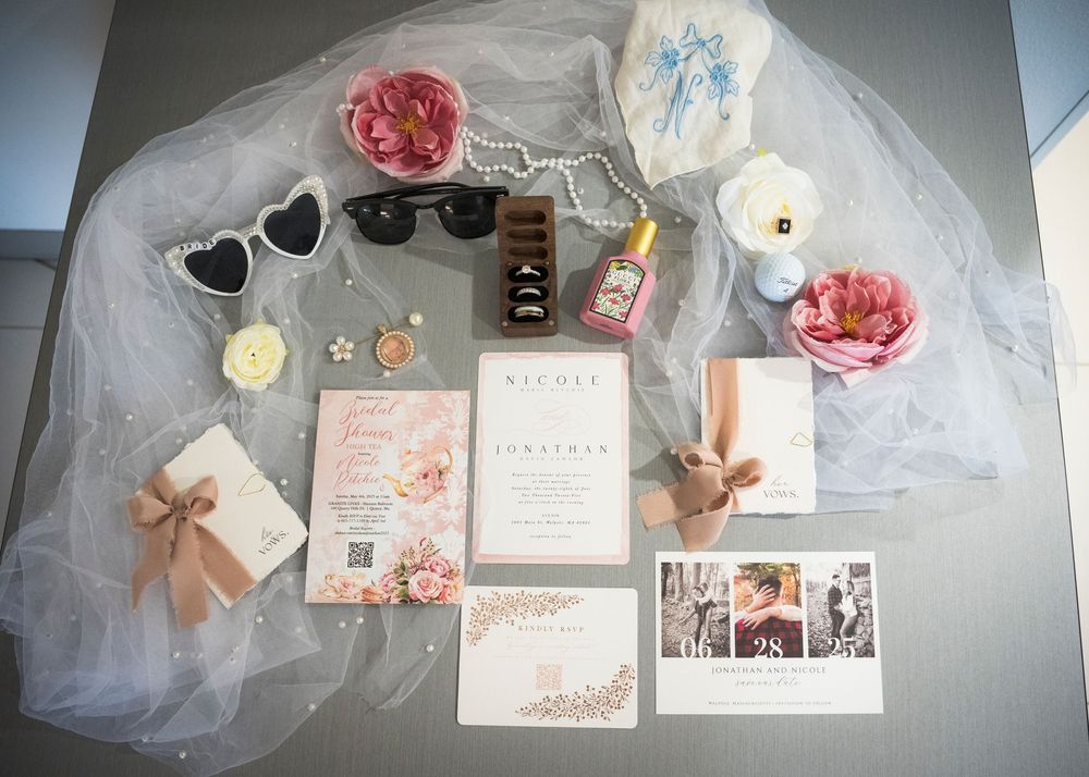 Flatlay of wedding invitations, heart sunglasses, rings, and floral details arranged on white tulle fabric.
