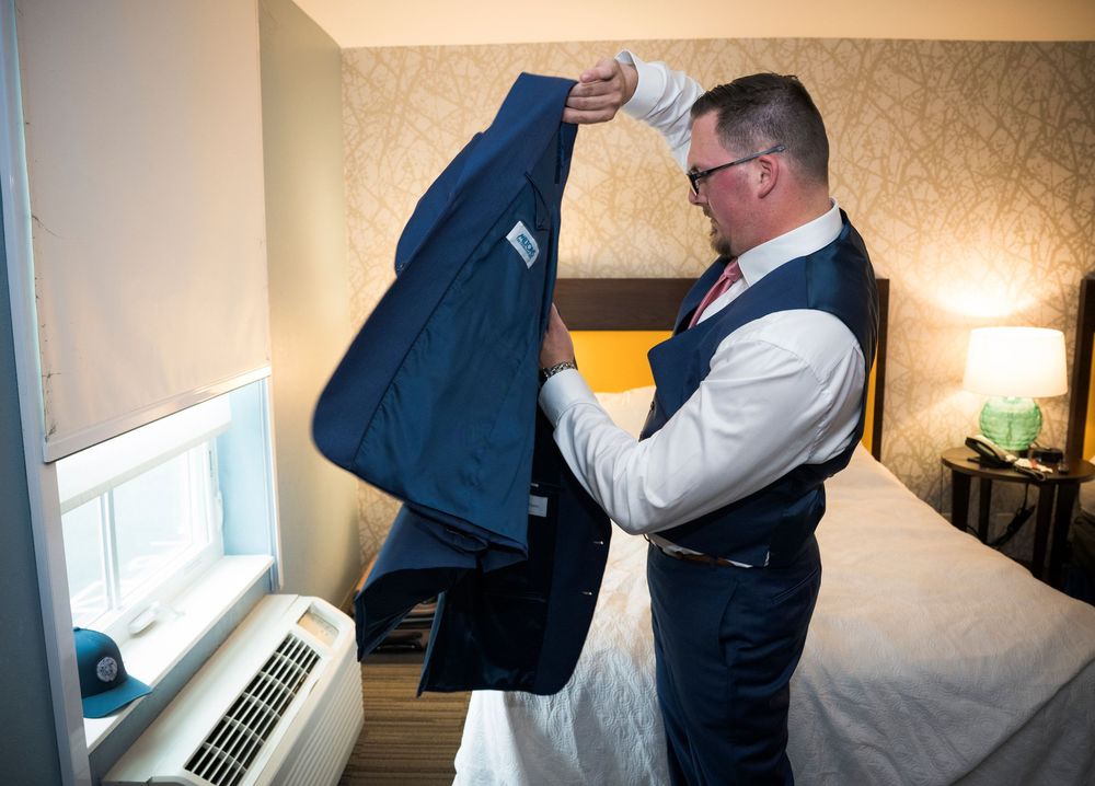 A person in formal attire examines a navy suit jacket while standing in a hotel room next to a window.