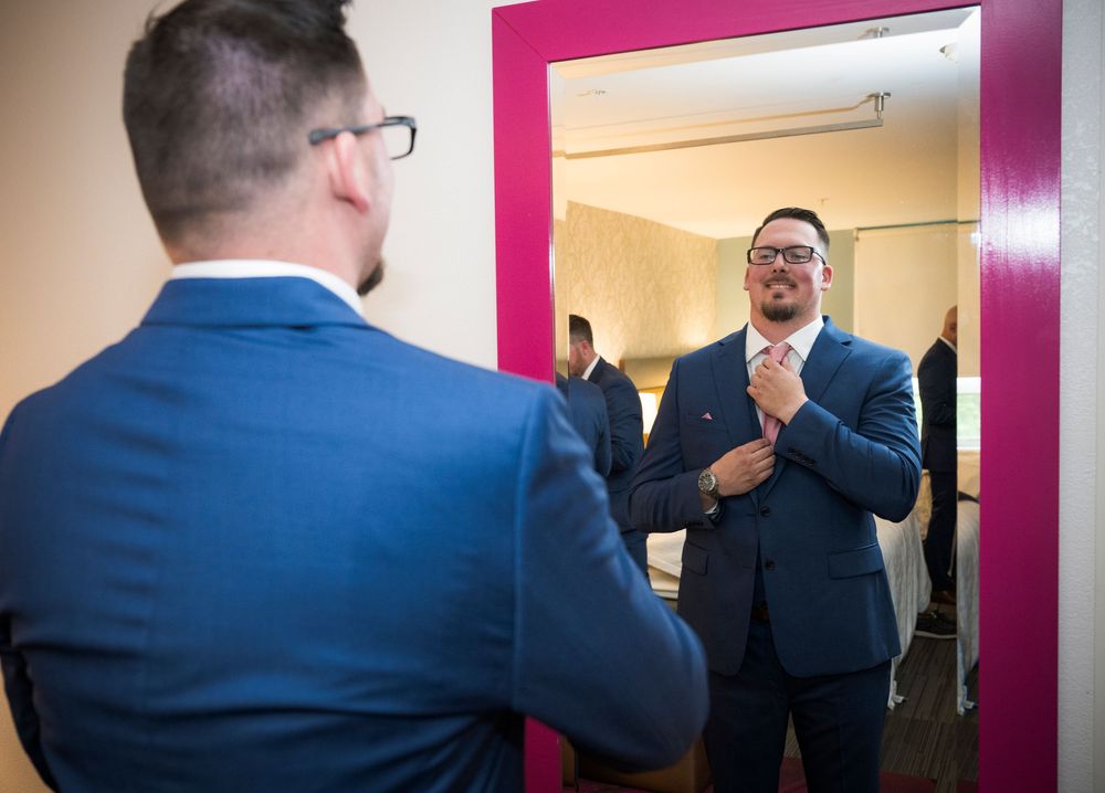 Reflection in pink-framed mirror shows person in blue suit adjusting tie while getting ready.