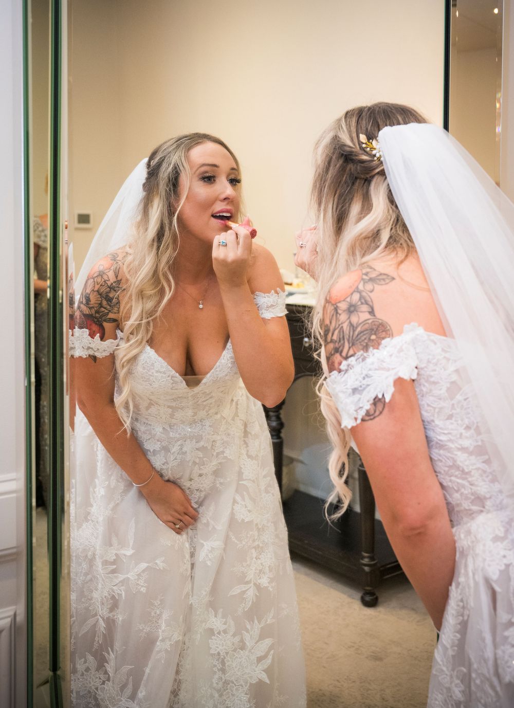 A bride wearing a white lace wedding dress with floral details looks at her reflection in a mirror while getting ready.
