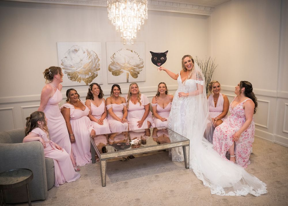 Wedding party in pink bridesmaid dresses gathered around bride in white gown under crystal chandelier in elegant room.