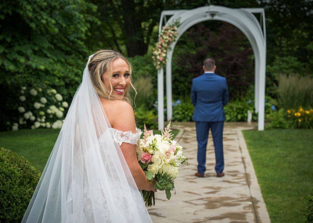 A wedding couple stands under a white garden arbor surrounded by lush summer greenery and flowers in an outdoor ceremony.