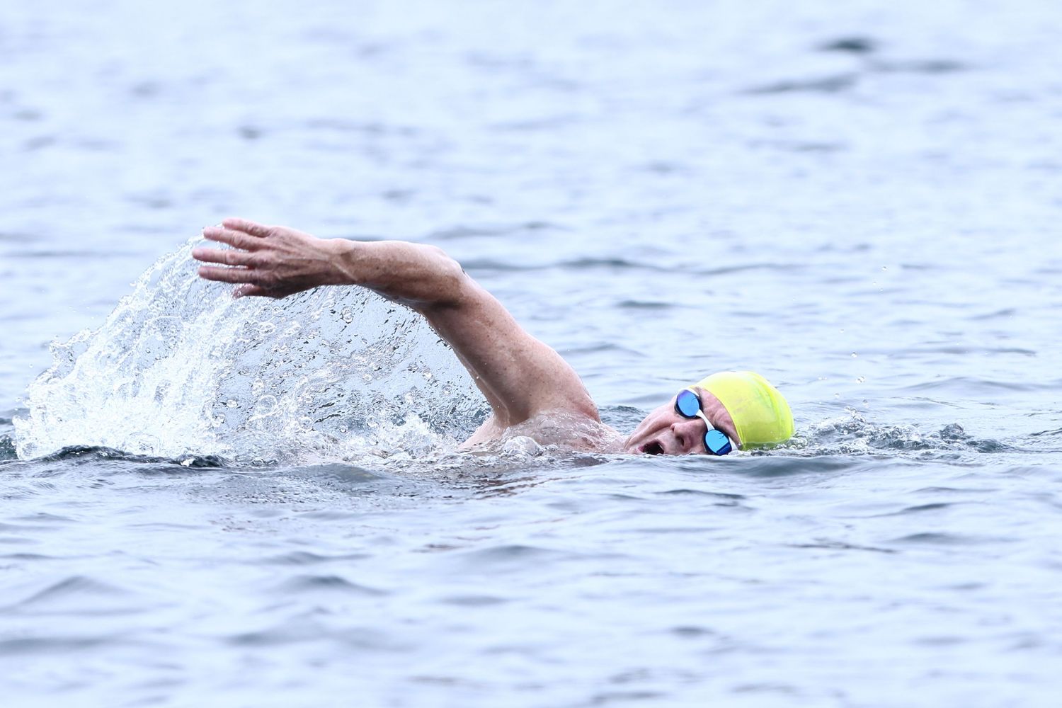 A swimmer performs freestyle stroke in open water wearing a yellow swim cap during a competition or training session.