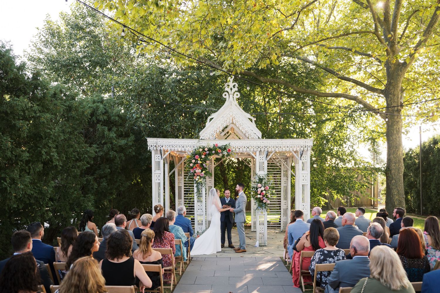 An outdoor wedding ceremony takes place under a white gazebo surrounded by trees and seated guests during golden hour.