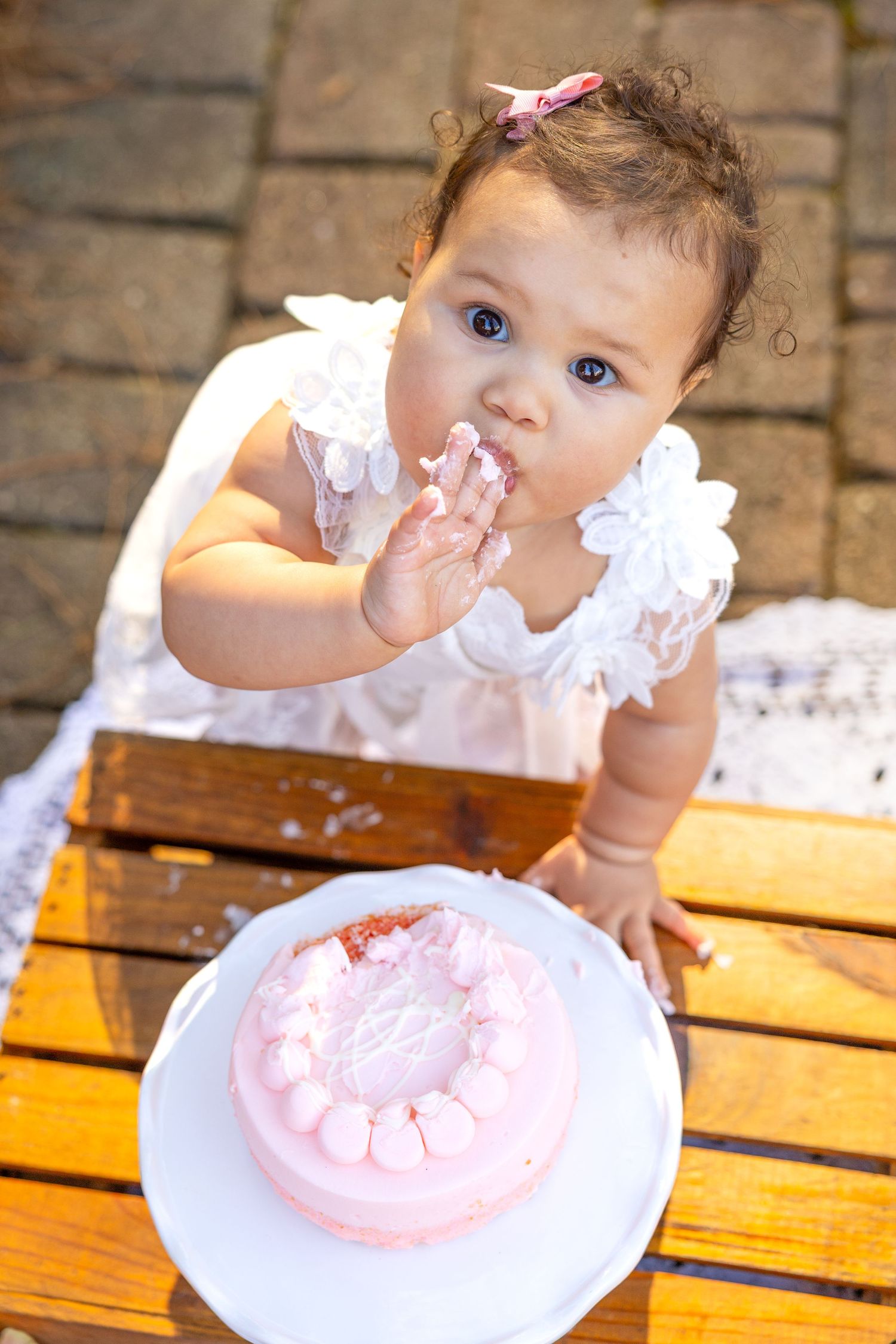 Baby in white dress enjoys first birthday cake with pink frosting while seated at wooden table outdoors at WRAL Azalea Gardens cake smash
