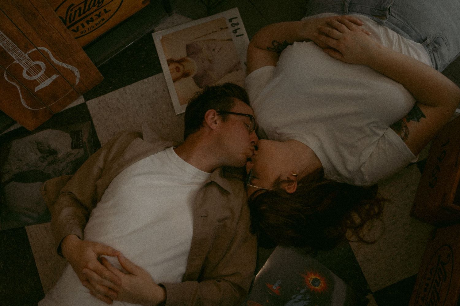 Two people in casual white clothes lying close together on the floor with scattered papers around them in dim lighting.