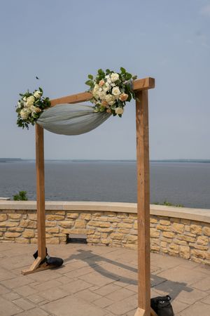 Wooden wedding arch decorated with white fabric and floral arrangements overlooking water.