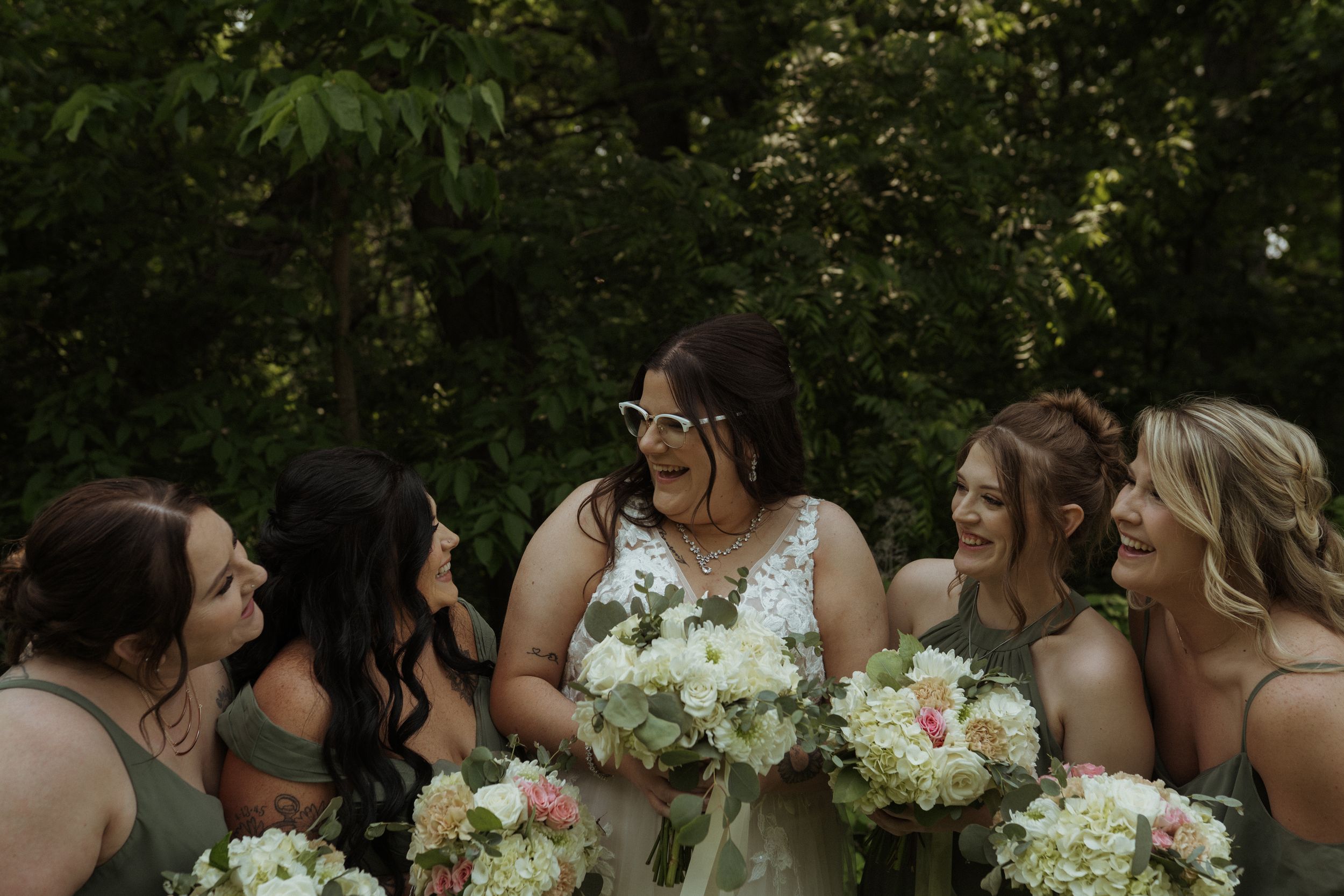 Bridesmaids in sage green dresses holding white bouquets pose outdoors.