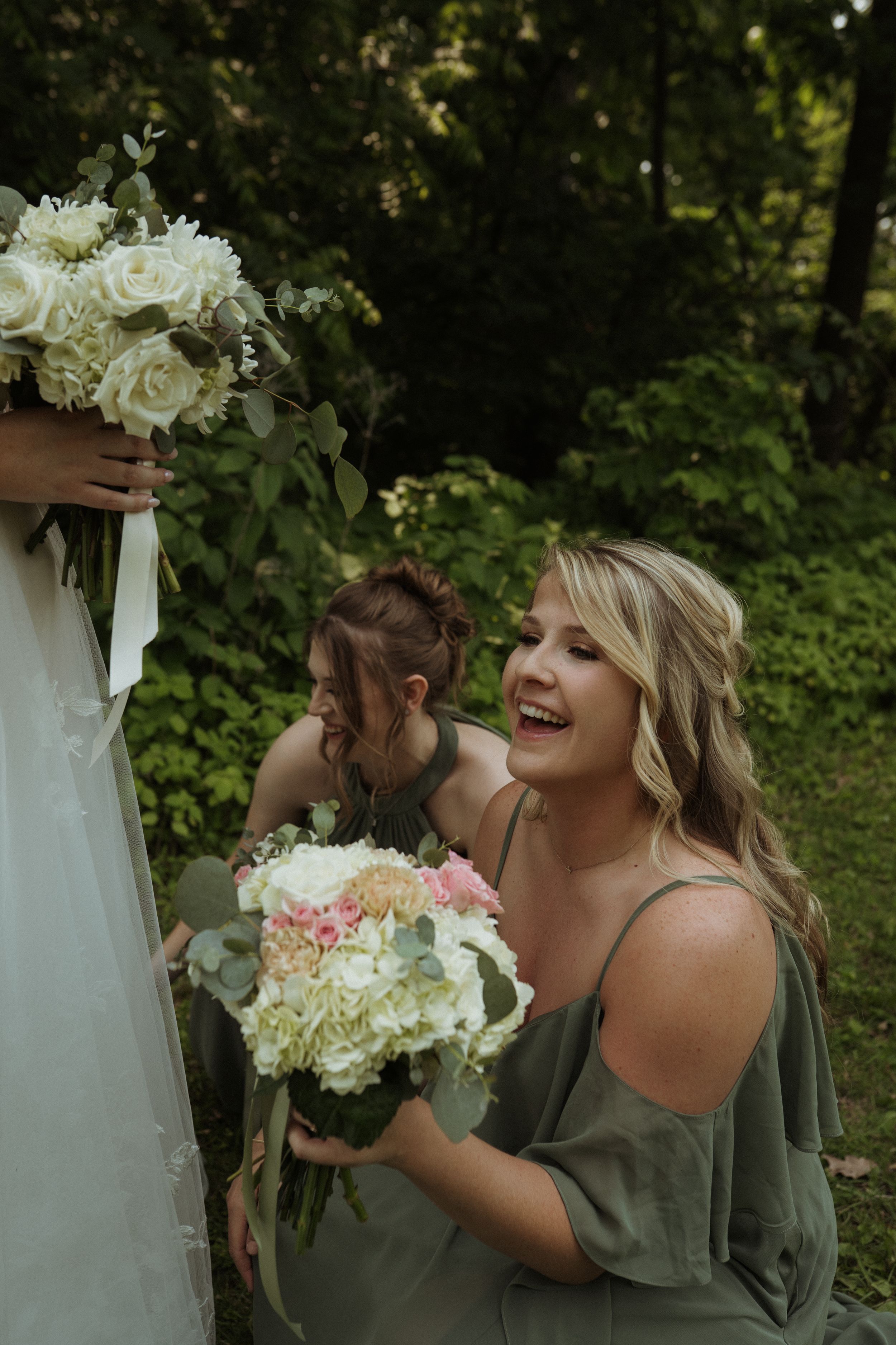 Bridesmaids in sage green dresses laugh while holding white and pink bouquets in an outdoor garden setting.