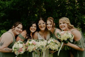 Wedding party with bridesmaids in sage green dresses holding bouquets smile together outdoors.