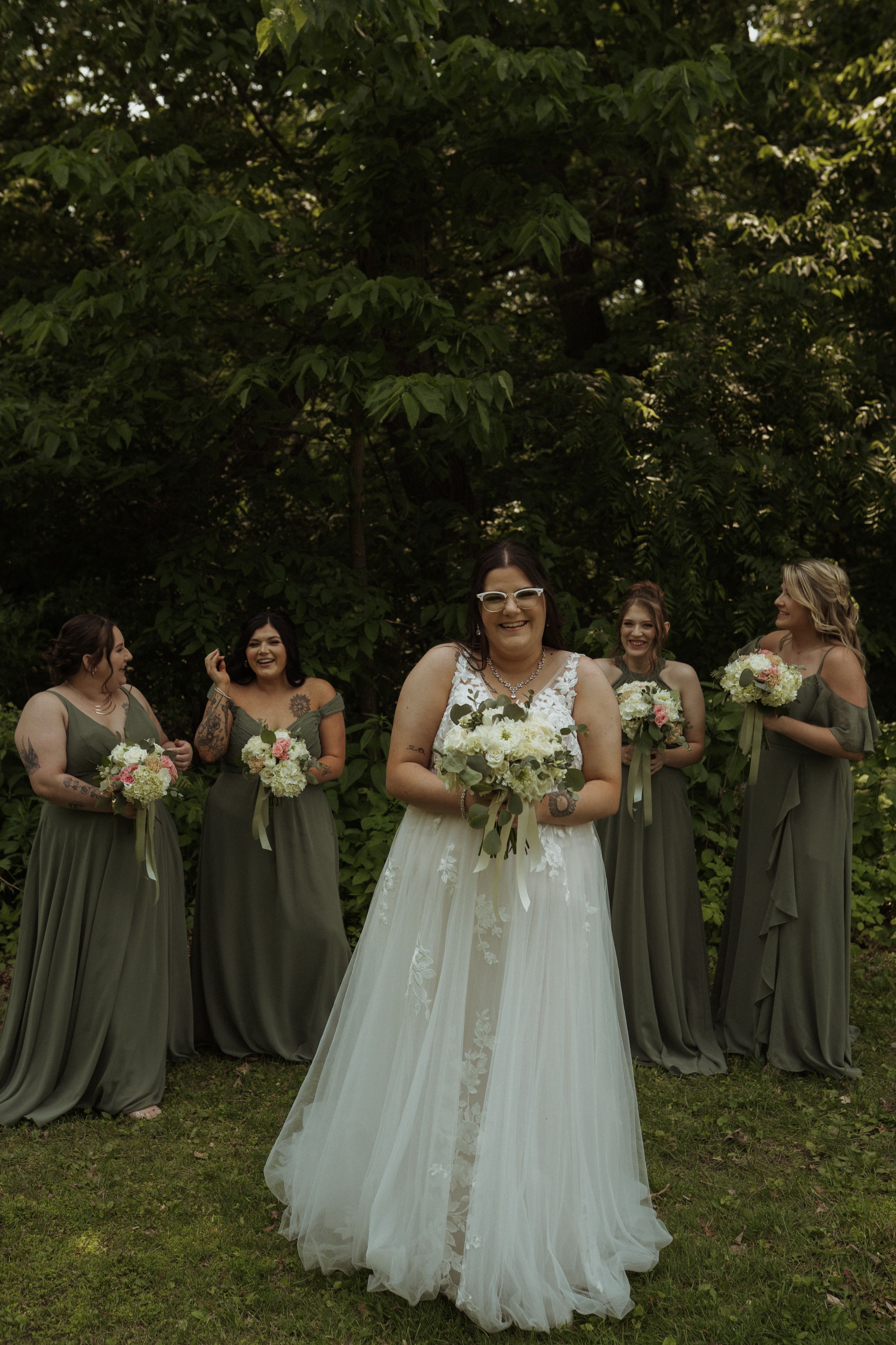 Bridal party in sage green dresses poses outdoors with bouquets and white-gowned bride.
