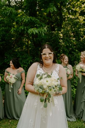 Bride in white lace gown poses with bridesmaids wearing sage green dresses in a garden setting.