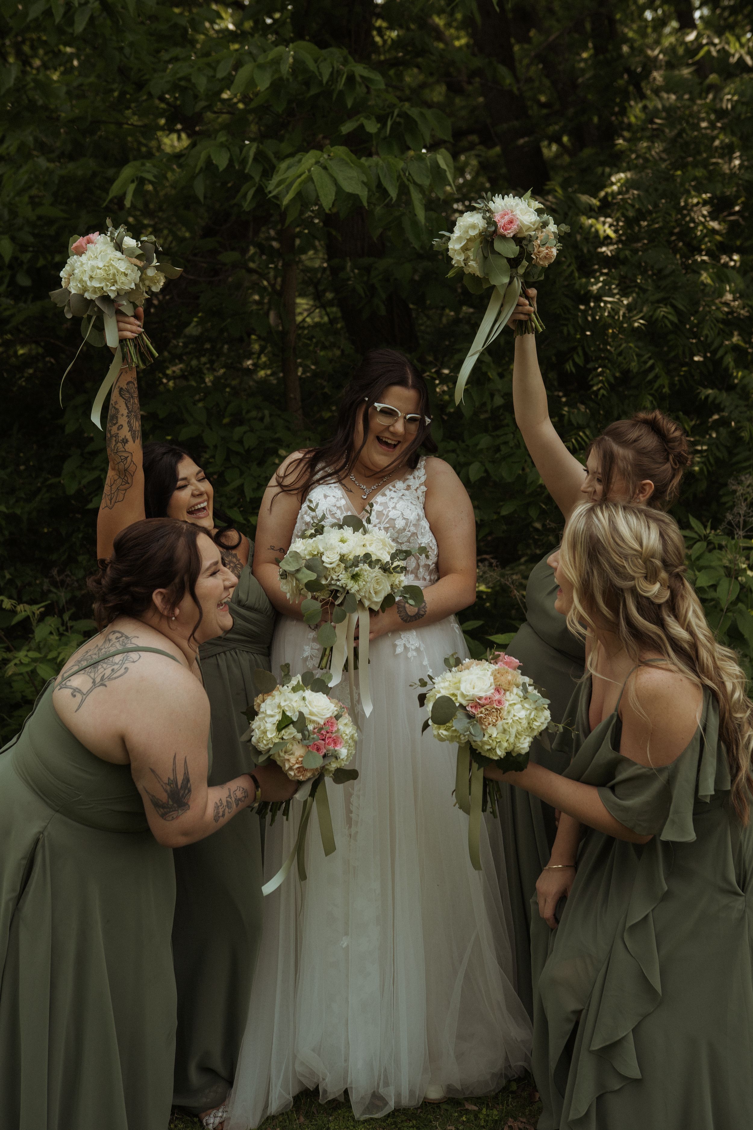Wedding party celebrates joyfully with raised bouquets surrounding bride in white gown against green foliage.