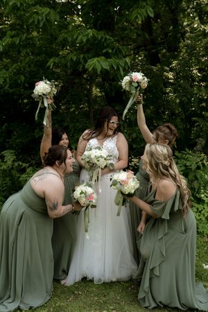 Bridesmaids in sage green dresses surround a bride holding white bouquets.
