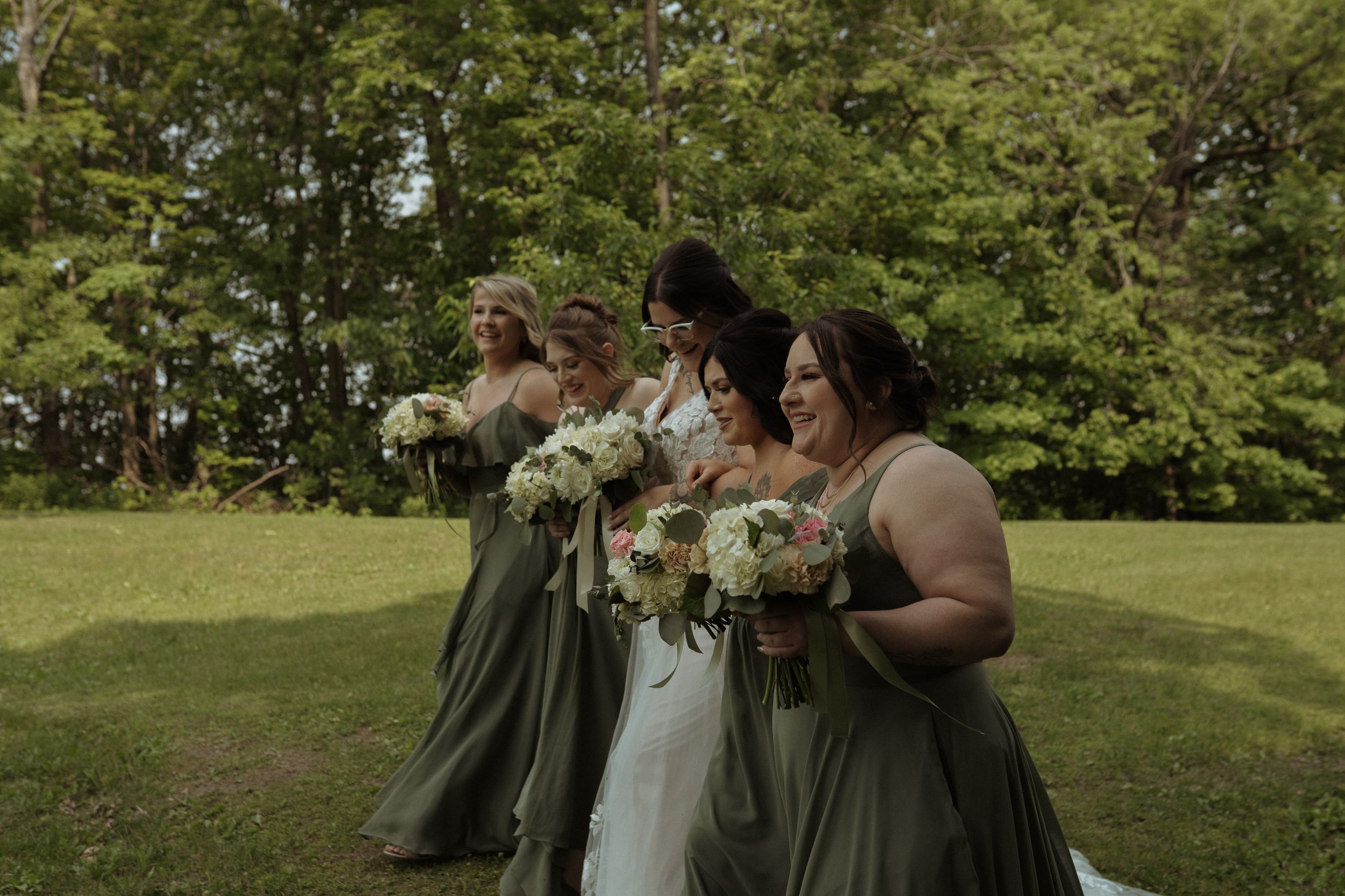 Wedding party in sage green dresses walks together holding bouquets on a sunny lawn.