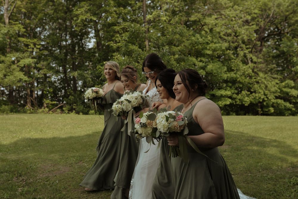 Wedding party in sage green dresses walks together holding bouquets on a sunny lawn.