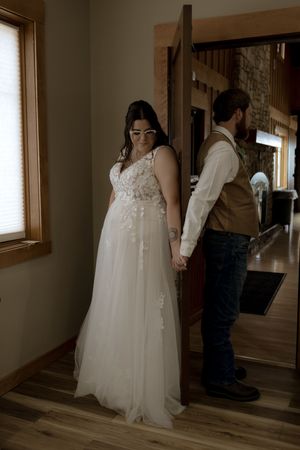 A couple holds hands while standing back-to-back in a rustic indoor setting before their wedding ceremony.
