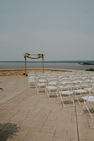 Outdoor waterfront wedding ceremony setup with white chairs and wooden arch on stone patio.
