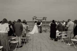 Black and white outdoor wedding ceremony with guests seated in folding chairs facing a scenic backdrop.