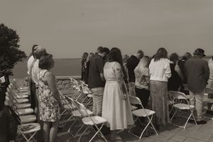 A vintage black and white photo shows people in formal attire standing near rows of folding chairs outdoors.