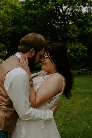 Candid wedding moment showing tender embrace on grassy field with green trees.