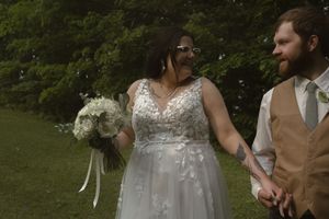 Wedding couple in white dress and tan vest embracing in a wooded outdoor setting.
