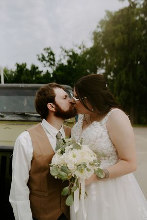 Romantic wedding kiss with white floral bouquet against a cloudy sky backdrop.