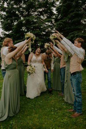 Wedding party in sage green dresses performs celebratory group toast on grassy lawn.