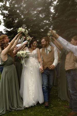 Joyful wedding group photo with bridal party in sage green dresses and tan vests.