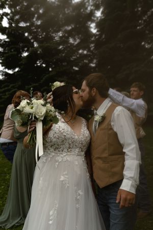 A couple shares a romantic kiss at their rustic outdoor wedding while holding a white floral bouquet.