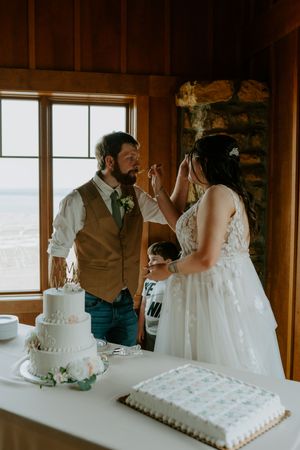 A playful cake-cutting moment at a wedding reception with a lake view through the window.