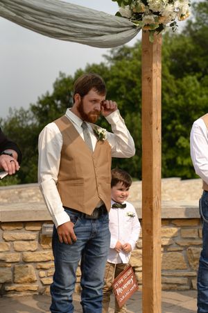 A groomsman in a tan vest and jeans stands under a wooden wedding arch decorated with white flowers.