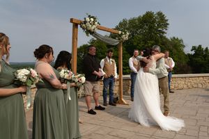 A wedding ceremony takes place under a wooden arch with bridesmaids in sage green dresses looking on.