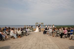 An outdoor wedding ceremony overlooking a lake with white chairs and wooden arch setup on stone patio.