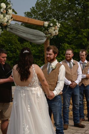 A wedding ceremony unfolds under a wooden arch adorned with white flowers and flowing fabric.
