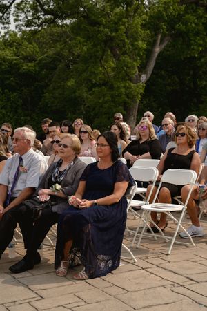 Wedding guests seated in white folding chairs watch the ceremony on a stone patio.
