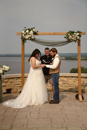 A wedding ceremony takes place beneath a wooden arch decorated with white flowers overlooking a lake.