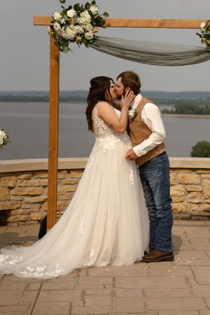 Newlyweds share their first kiss under a wooden arch decorated with white flowers and fabric draping.