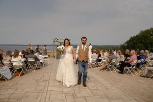 A wedding ceremony on a stone patio with guests seated in white chairs and a rustic wooden arch.