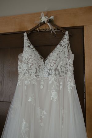 A delicate white lace wedding dress with floral appliques hangs on a wooden hanger.