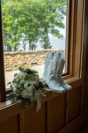 White cowboy boots and floral bouquet displayed on wooden windowsill with stone wall and trees visible outside.
