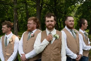 Groomsmen in tan vests and white shirts pose in line outdoors for wedding photos.