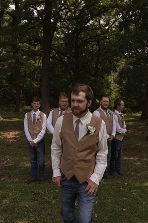 A wedding party in casual attire with tan vests stands in formation outdoors.