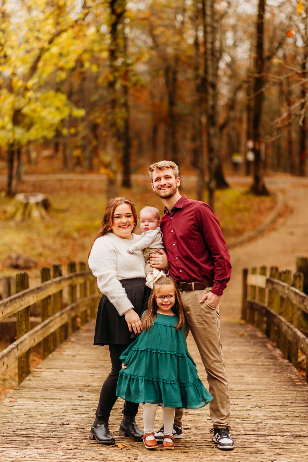 A family stands together on a wooden bridge in an autumn forest with golden foliage in the background.