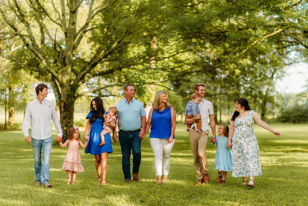 A group of people walking together through a sunlit park with green trees and grass on a warm summer day.