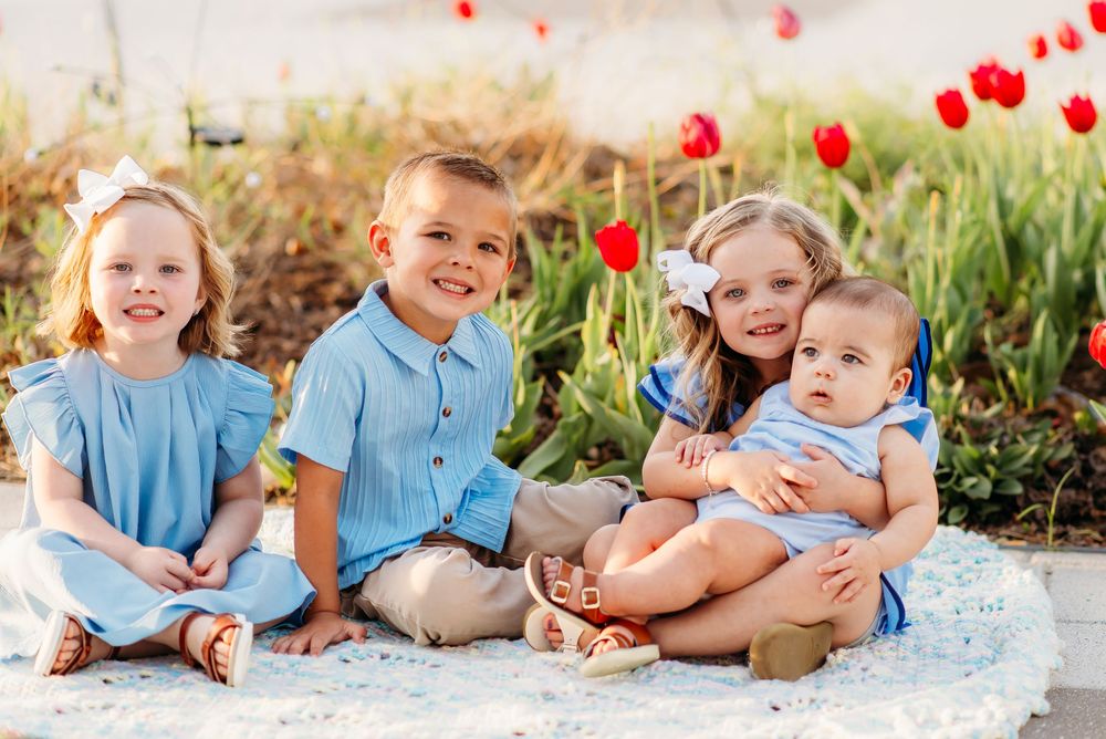 Four young children sit together on a blanket next to blooming red tulips in a spring garden setting.